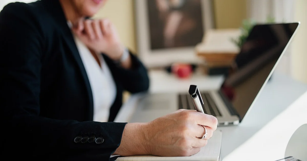 A business professional is seated at a sleek desk, actively taking notes with a pen while focused on a laptop screen. Natural light enhances the workspace's ambiance.